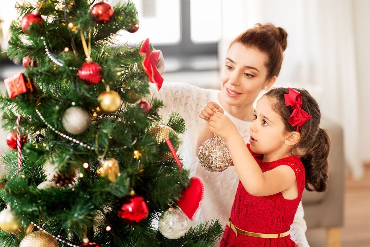 A woman and a young girl partake in beloved holiday traditions as they decorate a Christmas tree together. The girl, dressed in a red dress with a matching bow, hangs a gold ornament on the tree, which is adorned with red and gold decorations. Both appear focused and content.