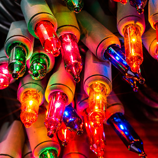 Close-up of colorful holiday string lights glowing vividly in green, red, blue, and orange. The lights have a reflective, glossy finish, with gray bases and are tightly packed together, creating a festive and vibrant display.