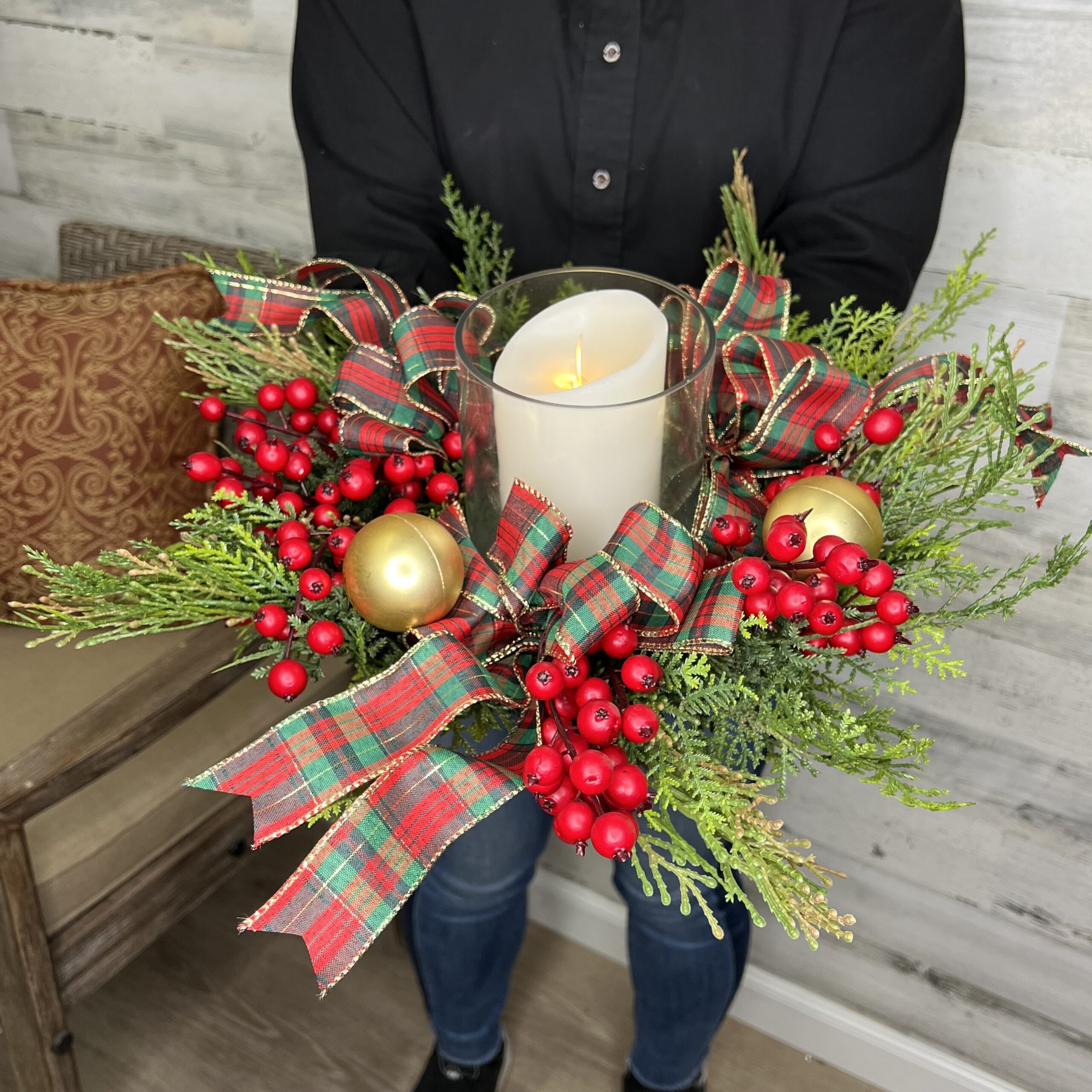 A person holds a festive arrangement featuring a white candle in a glass holder, surrounded by red berries, green foliage, plaid ribbons, and gold ornaments. The person wears a black shirt and jeans, standing indoors on a wooden floor.