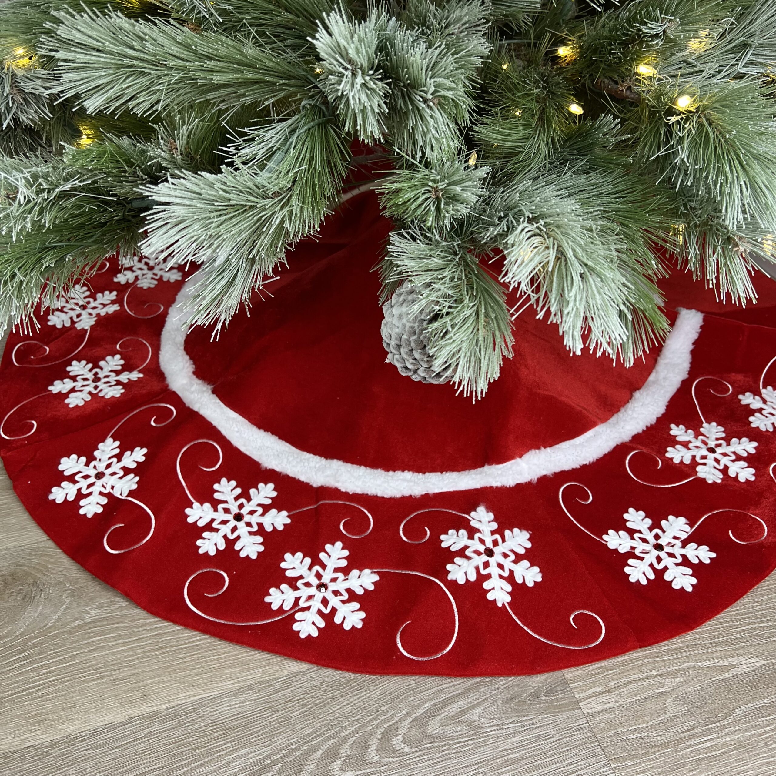 Red Christmas tree skirt with white snowflake designs and swirls beneath a decorated tree with frosted branches and yellow lights. The skirt features a white fur trim. The tree stands on a wooden floor.