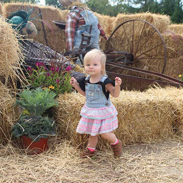 A toddler wearing denim overalls and a pink-patterned dress walks on straw, surrounded by hay bales. Behind her, there are rustic decorations, including a scarecrow and a wagon wheel, along with a potted plant.