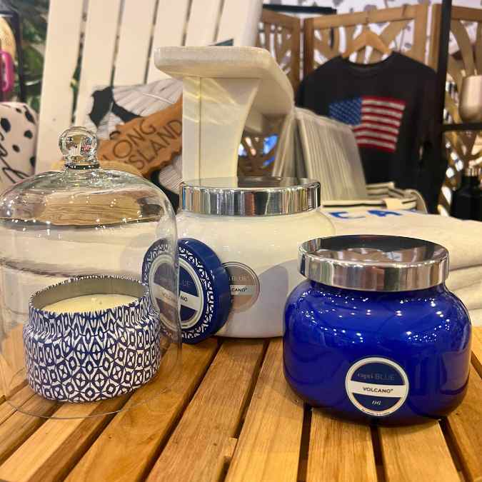 Several decorative candles in blue and white jars, one under a glass cloche, displayed on a wooden table with textiles and pillows in the background.