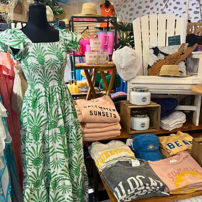 A display in a boutique features a green and white dress on a mannequin, folded graphic t-shirts, hats, mugs, stacked towels, a wooden chair, and beach-themed decor items, creating a summer vibe.