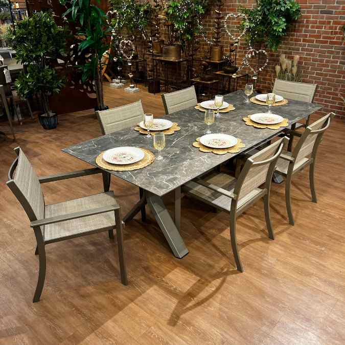 A rectangular stone-top dining table with six beige chairs is set with plates, glasses, and gold chargers. The table sits on a wooden floor, surrounded by plants and a brick wall in the background.