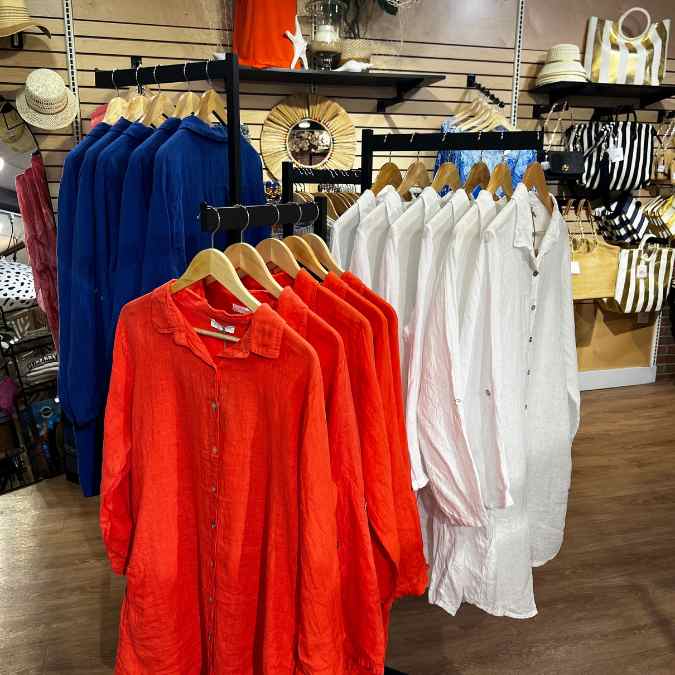 A display at Hicks Nurseries new shop features long-sleeved shirts in red, blue, and white on wooden hangers. In the background, bags and hats are displayed on shelves and hooks.