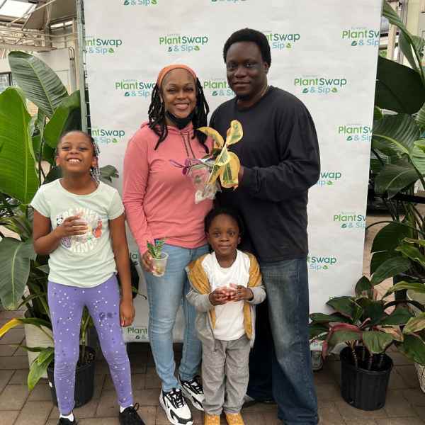 A smiling family of four stands together at a Plant Swap & Sip event, holding small potted plants. They are surrounded by green plants and a branded backdrop.