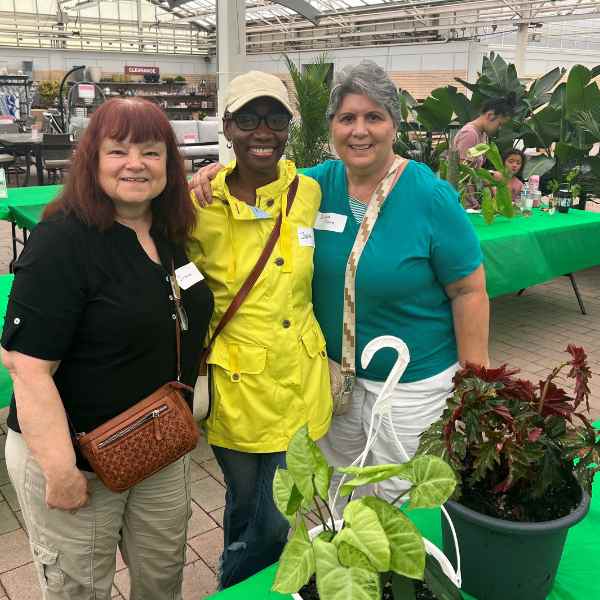 Three women smile and pose together indoors at a plant workshop, standing behind a table with potted plants. The setting appears to be a greenhouse, with green tablecloths and large leafy plants in the background.