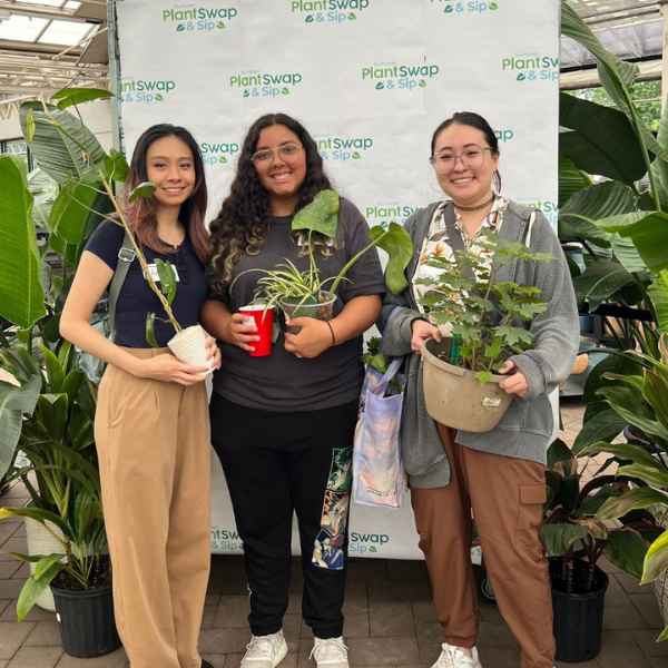 Three women stand smiling in front of a Plant Swap & Sip banner, each holding a potted plant. Tall green plants surround them, and they appear to be at a plant-themed event or gathering.