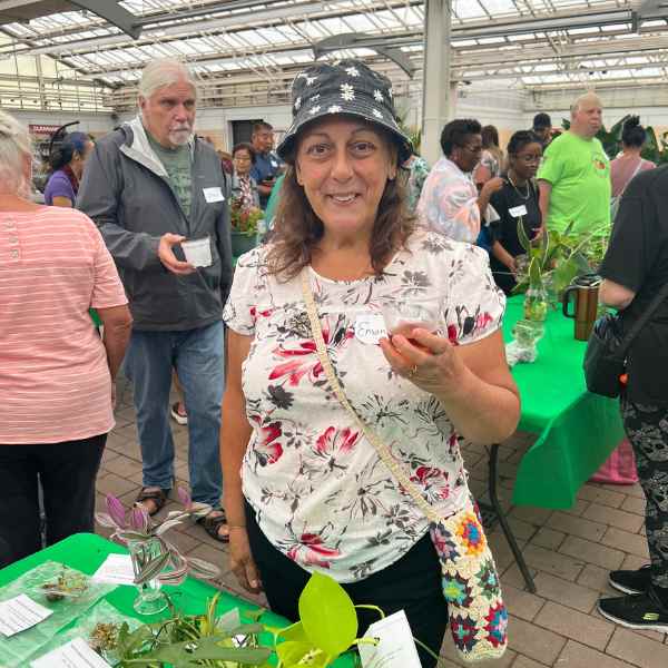 A woman in a floral shirt and flower-patterned bucket hat smiles and holds a small plant at a busy indoor plant event; people and green tables with plants are visible in the background.