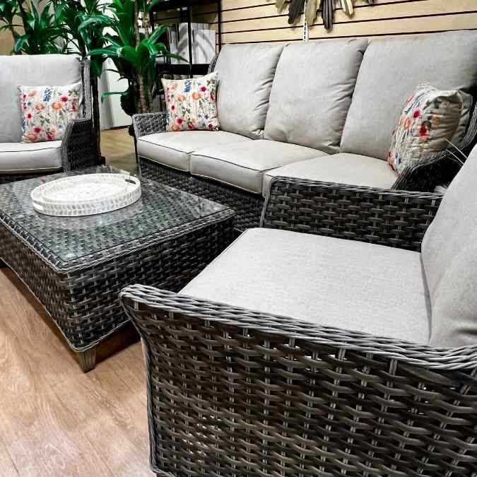 Wicker patio furniture set with light beige cushions, two armchairs, a sofa, a glass-topped coffee table, and floral accent pillows. Wooden floor and green plants in the background.