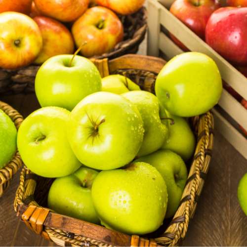Apples A wicker basket filled with green apples sits on a wooden table, surrounded by crates of red and yellow apples in the background.