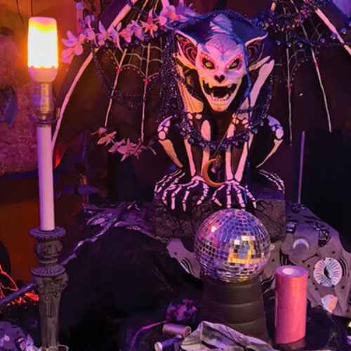 Hicks Nurseries Fall Animated Story featuring a skeleton, disco ball and light A spooky, winged gargoyle with glowing red eyes sits behind a disco ball and candles, surrounded by dark fabrics, fake cobwebs, and eerie purple lighting.