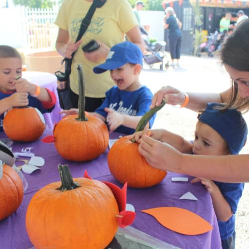 Hicks Nurseries Fall Festival (1) Four young children and an adult decorate pumpkins with paper shapes at a table covered in a purple cloth, enjoying a festive outdoor activity. The kids appear focused and excited.