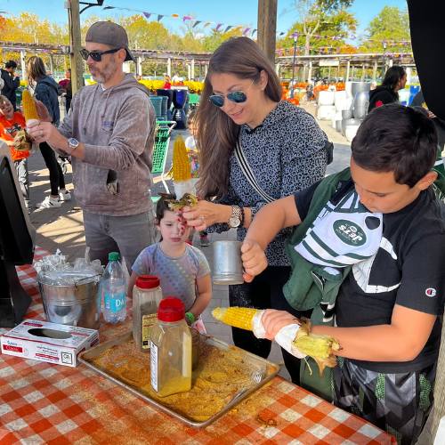 Hicks Nurseries Fall Festival (11) A woman and two children prepare corn on the cob at an outdoor market table covered with spices and toppings, while a man stands nearby. Other people and pumpkins can be seen in the background.