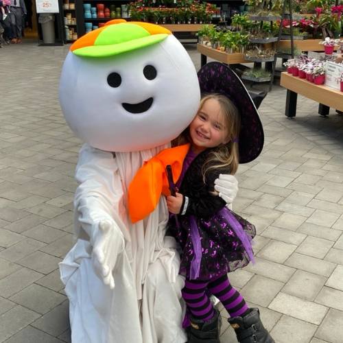 Hicks Nurseries Fall Festival (4) A smiling child in a purple witch costume hugs a large person in a happy ghost costume at a garden center, surrounded by plants and flowers on display tables.