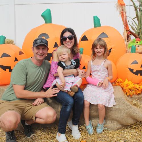 Hicks Nurseries Fall Festival (5) A family of four smiles while posing in front of large inflatable jack-o’-lanterns at a pumpkin patch. The parents sit with their two young daughters, surrounded by autumn decorations and hay.