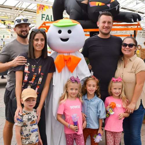 Hicks Nurseries Fall Festival Activities (2) A group of six people, including three adults and three children, pose and smile with a person in a ghost costume at a fall-themed event. Decorations and a large inflatable cat are visible in the background.