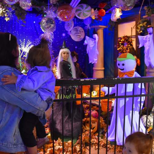 Hicks Nurseries Fall Festival Walkthru Animation A parent holds a child while viewing a festive Halloween display with witches, ghosts, pumpkins, and a snowman figure, all surrounded by colorful decorations and autumn leaves. Another child stands nearby.