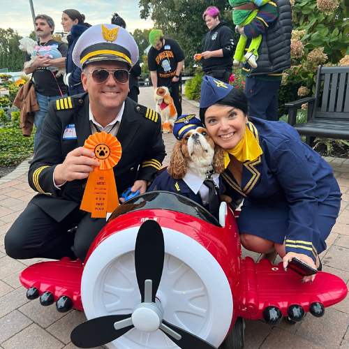 Hicks Nurseries Pet Parade (2) Two adults dressed as pilots and a dog in a pilot outfit pose with a red toy airplane. The man holds an orange ribbon. People and dogs in costumes are in the background at an outdoor event.