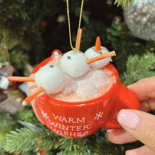 A close-up of a red mug ornament with Warm Winter Wishes written on it, filled with marshmallow snowmen melting in frothy hot chocolate, held in front of a Christmas tree.