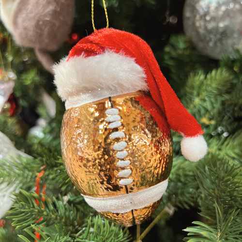 A gold football-shaped ornament with white laces is hanging on a Christmas tree and is topped with a small red and white Santa hat.
