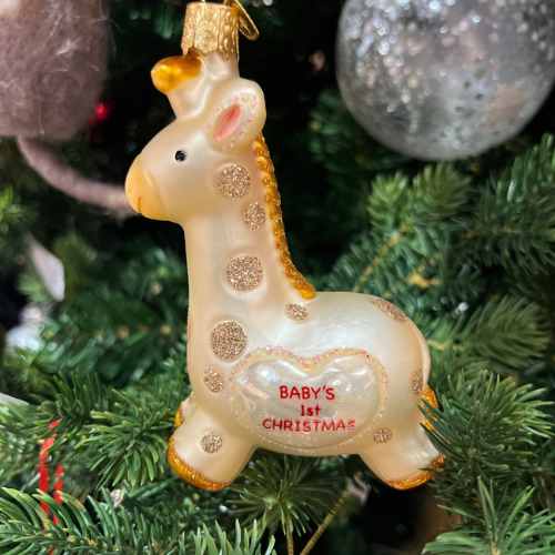 A giraffe-shaped Christmas ornament with gold accents hangs on a tree. The ornament has BABYS 1st CHRISTMAS written in red letters on its side. In the background, there are green branches and other decorations.