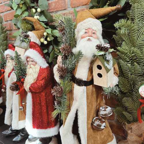 Christmas Decor Santas Four Santa Claus figurines are displayed by evergreen branches; one in a gold robe holds a birdhouse and pine branches, while the others wear traditional red suits with white trim.
