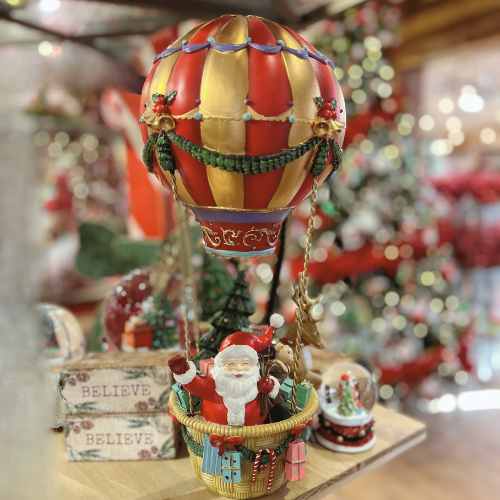 Christmas Decor Santa in Hot Air Balloon A festive figurine of Santa Claus riding in a hot air balloon basket decorated with gifts, with a red and gold balloon above him. Christmas trees and ornaments are blurred in the background.