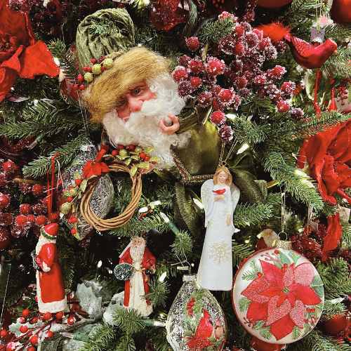 A Christmas tree decorated with a Santa Claus face ornament, red berries, red flowers, a white angel, Santa figurines, and a round ornament with a red poinsettia design among green branches.