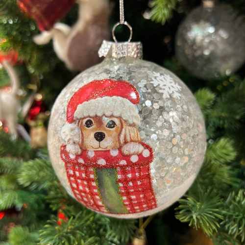 A Christmas tree ornament featuring a cute dog wearing a red Santa hat and peeking out of a red-and-white checkered stocking, surrounded by glittery snowflake patterns.