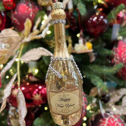 A glittery champagne bottle ornament with a Happy New Year label hangs on a Christmas tree decorated with red baubles and golden leaves.