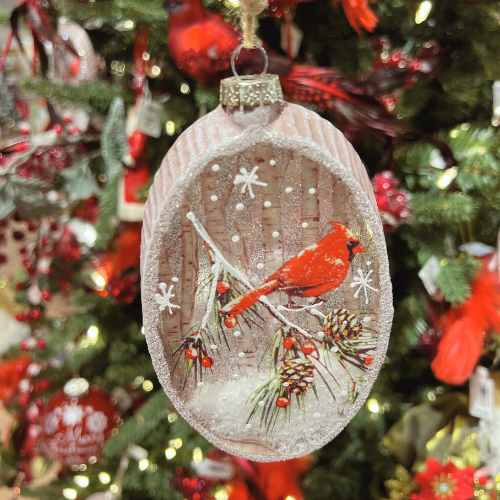 An oval Christmas ornament featuring a red cardinal perched on a snowy pine branch, decorated with pinecones and red berries, hangs on a Christmas tree with twinkling lights and festive decorations in the background.