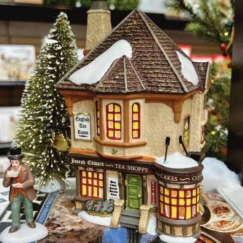Dept 56 Villages Collectibles (1) A miniature English tea shop model decorated for Christmas with snow on the roof, a Christmas tree beside it, and a figurine of a man in Victorian attire holding a cup.
