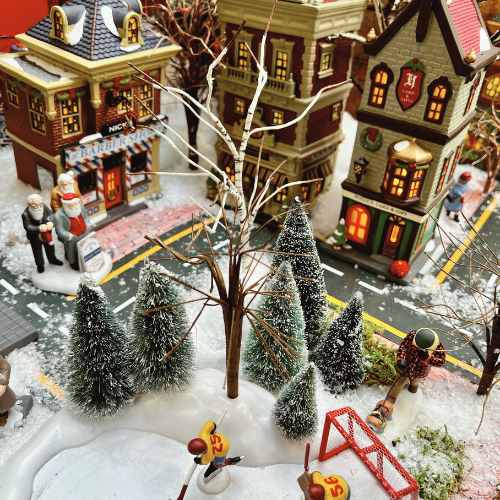 Dept 56 Villages Collectibles (2) A festive miniature winter village scene with snow-covered trees, people, and colorful buildings. A Santa figure stands near a shop, and a child plays on a red swing set in the snow-covered park.