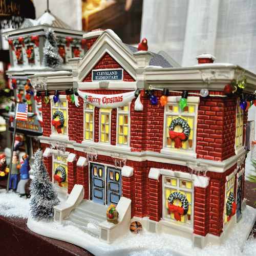 Dept 56 Villages Collectibles (4) A miniature model of a red brick building labeled Cleveland Elementary decorated with wreaths, lights, and a Merry Christmas sign, with snowy details and tiny festive figures outside.