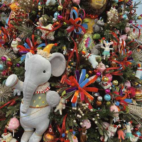 A Christmas tree decorated with colorful ornaments, ribbons, and plush toys, including a large gray elephant wearing a hat and circus outfit, with various animal figures and baubles throughout the branches.