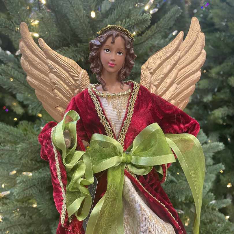 A Christmas angel tree topper with golden wings, curly brown hair, and a red velvet robe tied with a large green ribbon, stands in front of a decorated Christmas tree.