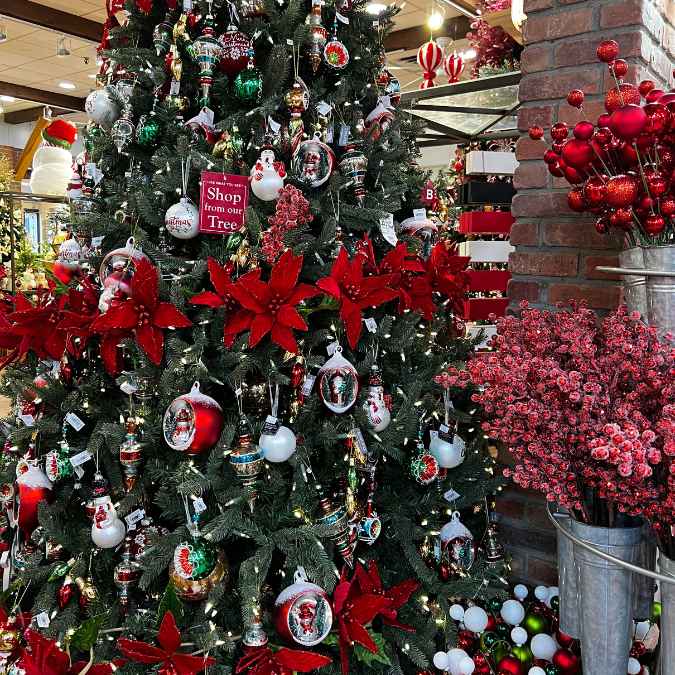 A Christmas tree decorated with red poinsettias, ornaments, and snowmen stands beside metal buckets filled with red berry branches in a festive store display. A sign on the tree reads, Shop from our Tree.