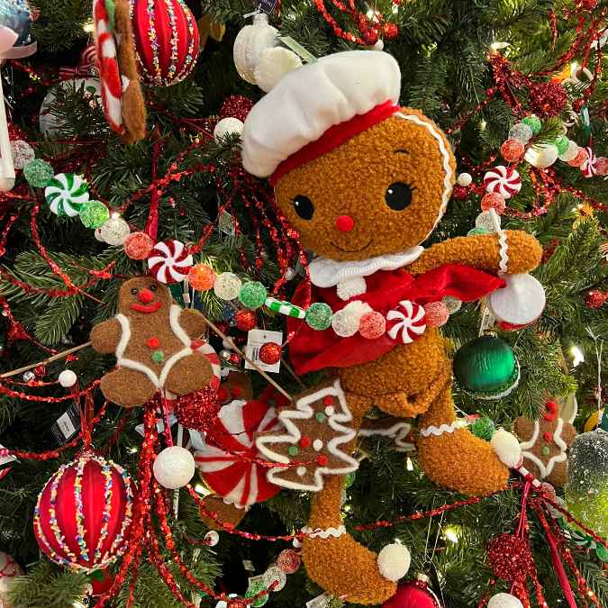 A gingerbread doll with a white chef hat and red scarf hangs on a Christmas tree decorated with candy ornaments, gingerbread cookies, striped garlands, and colorful baubles.