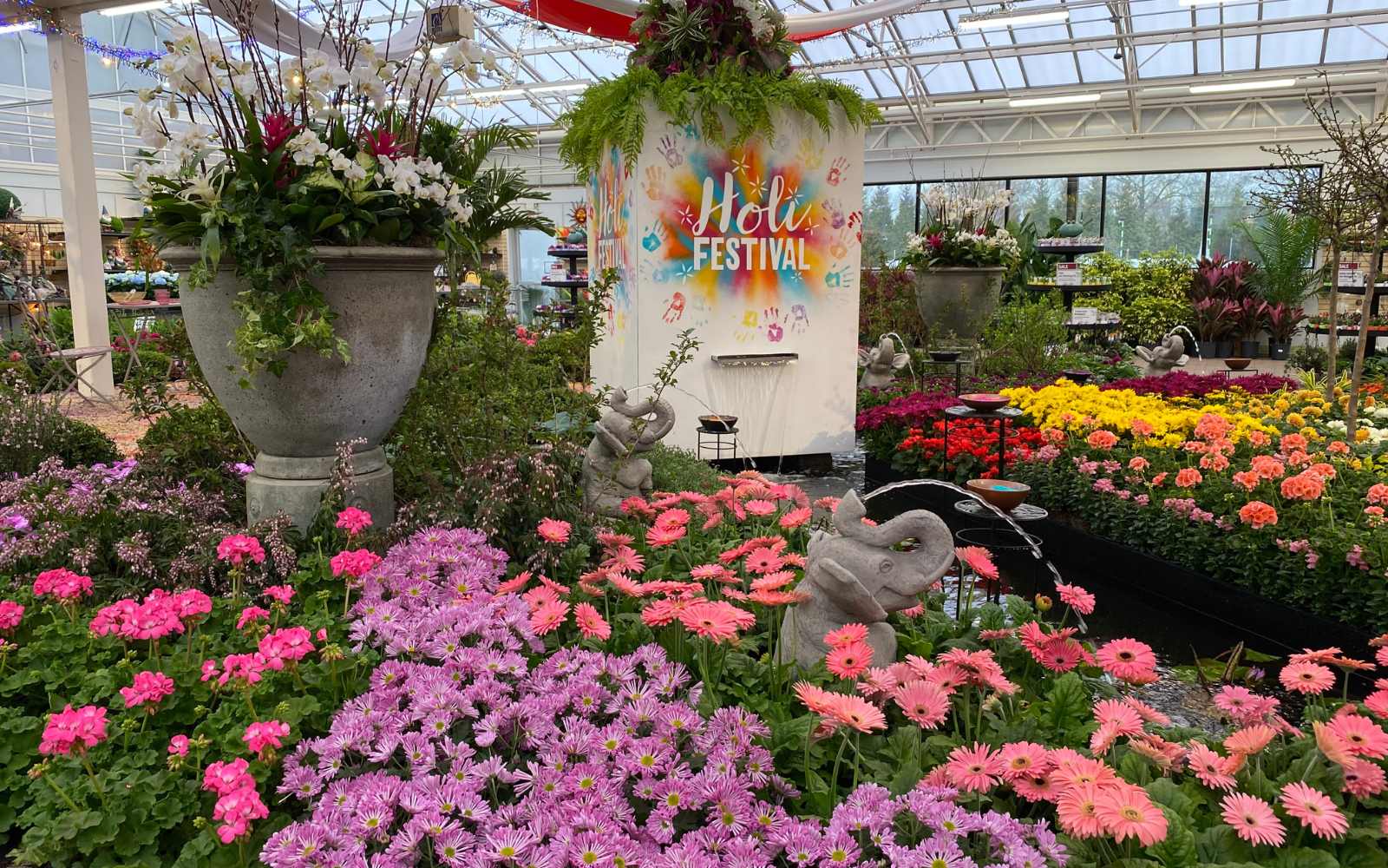 A vibrant indoor garden with colorful flowers, potted plants, elephant statues, and a “Holi Festival” sign in the background, under a glass ceiling with natural light.