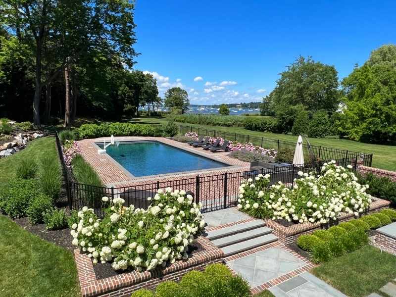 A rectangular swimming pool is surrounded by a brick patio, black fence, and blooming white flowers. Lounge chairs and umbrellas sit nearby, with green lawns, trees, and a view of water in the distance under a blue sky.