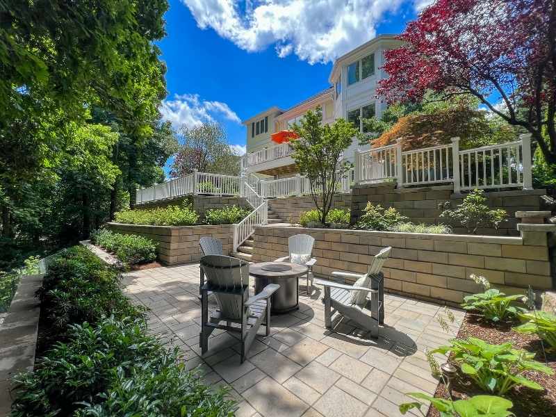 A sunlit patio with four Adirondack chairs around a fire pit, surrounded by garden beds and terraced stone retaining walls, in front of a large white house with multiple decks and lush landscaping.