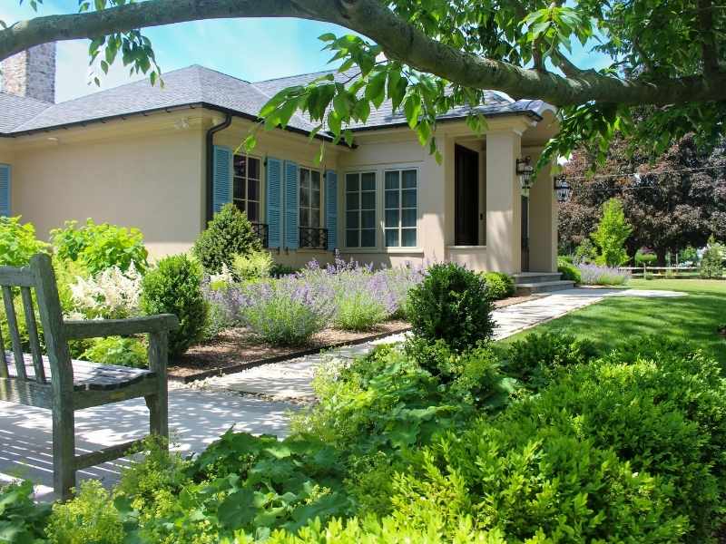 A single-story beige house with blue shutters, surrounded by lush green shrubs, flowering plants, and a well-kept garden. Sunlight filters through a tree, and a wooden bench is in the foreground.