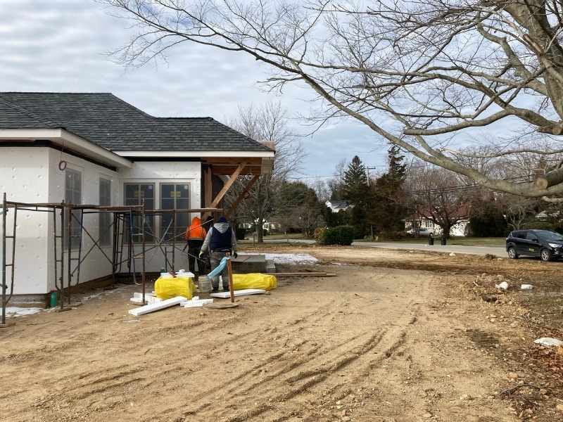 Two construction workers stand next to scaffolding and building materials beside a partially constructed house. The yard is bare dirt, and a car is parked near leafless trees on the right side of the image.