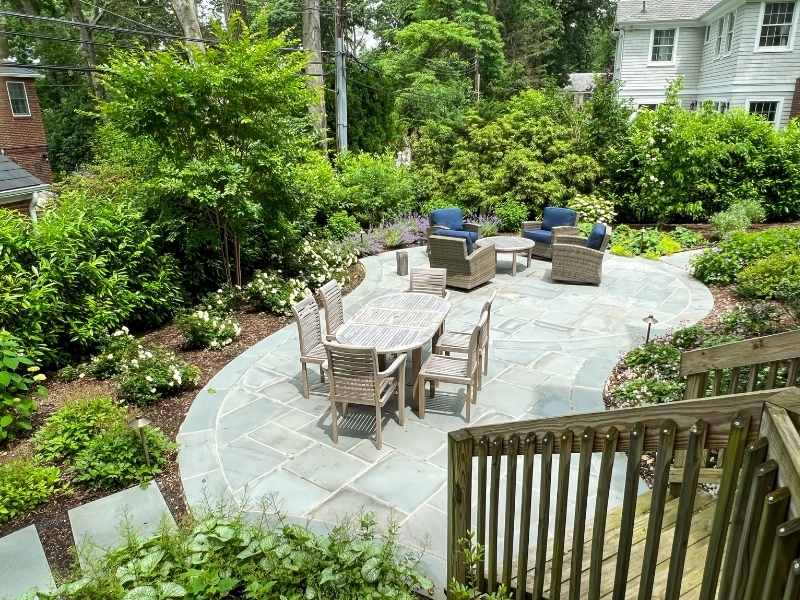 A backyard patio with a circular stone floor features a dining table with wooden chairs and a separate seating area with blue cushioned chairs, surrounded by lush green plants and flowers.