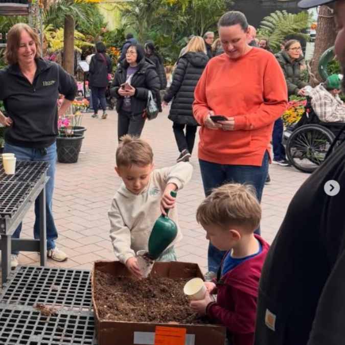 Two young boys scoop soil into cups at an outdoor gardening event, while adults and other visitors watch and walk around in a busy, plant-filled area.