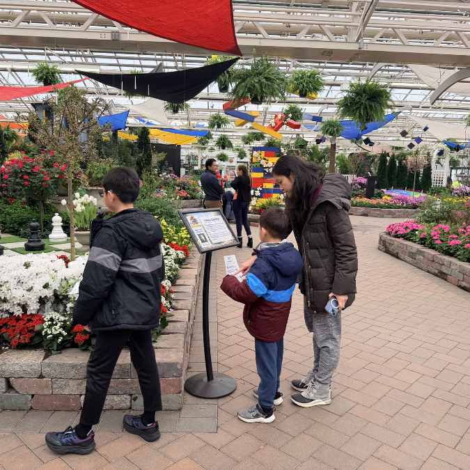 Three children in jackets read an informational display in a bright indoor garden with colorful flowers, hanging plants, and geometric fabric decorations overhead. Other visitors walk in the background.