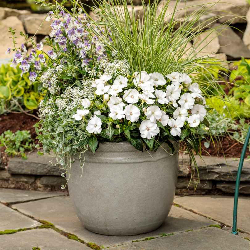 A gray ceramic planter filled with white flowers, tall ornamental grass, and greenery sits on a stone patio, with a garden and stone wall in the background.