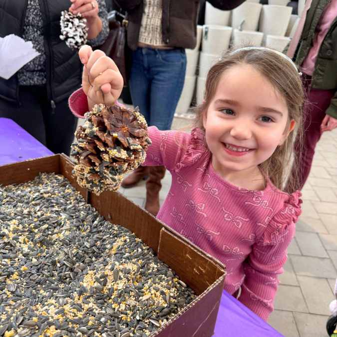 A smiling young girl holds up a pinecone covered in birdseed at a craft table, where a box is filled with seeds. People and cinder blocks are visible in the background.