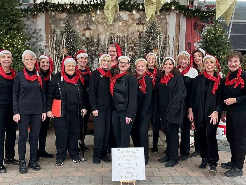A group of women dressed in black outfits and red scarves, wearing Santa hats, stand smiling in front of decorated trees and holiday lights. A festive sign reading Happy Holidays is displayed in front of them.